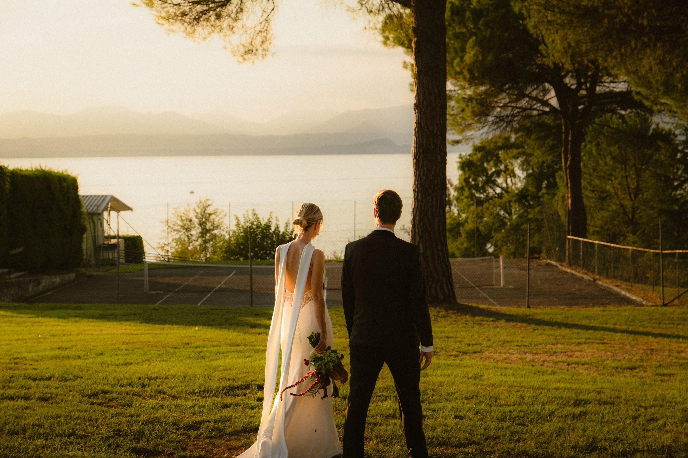 Braut und Bräutigam von hinten auf der Wiese, vor ihnen der Gardasee im Abendlicht
