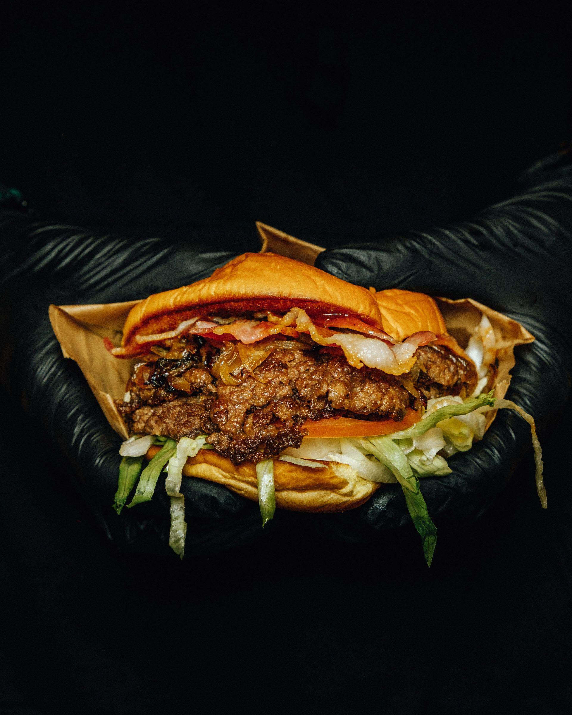 Fertiger Doppel-Burger mit Bacon und Cheddar in einer behandschuhten Hand vor schwarzem Hintergrund